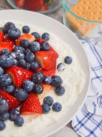 Ready to serve up this delicious and patriotic dessert! It has been on my table for years for the 4th of July or Memorial Day! Everyone loves it and I love how beautiful it is. You can even lay out your fruit to make a flag design.  How to Make Patriotic Cheesecake Dip: This recipe is so simple, let's start by adding your cream cheese to a stand mixer or mixing bowl if using a hand mixer. Whip together until nice and creamy. Add greek yogurt and vanilla extract and mix together. Next, add your powder sugar. Mix together once more until well combined and creamy. Remove from mixer and add to a serving bowl. Top fresh strawberries and blueberries. Serve graham crackers for dipping!