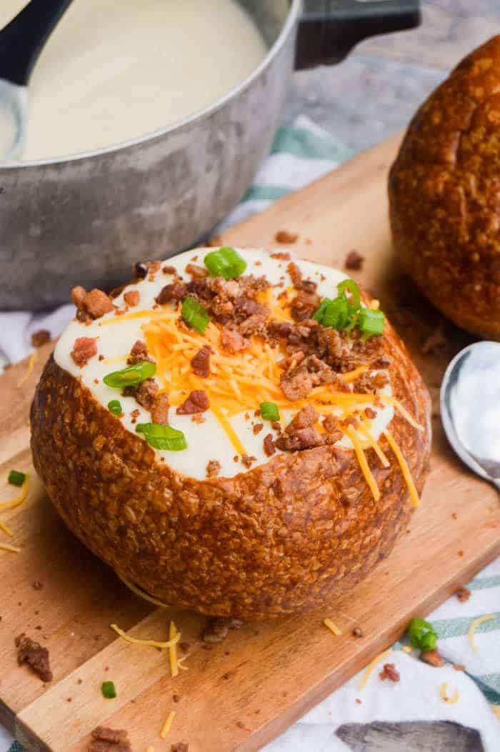 Potato soup in a bread bowl topped with cheese, bacon bits, and chives on a wooden board