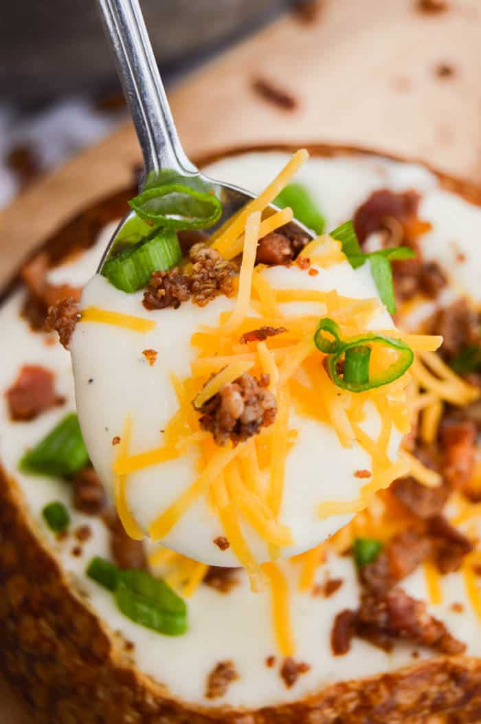 Close up of spoonful of potato soup above a bread bowl, topped with cheese, bacon, and chives