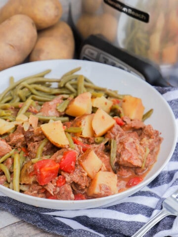 Slow Cooker Steak and Potatoes in a white bowl with slow cooker and potatoes in back