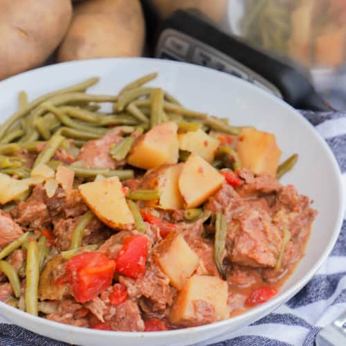 Slow Cooker Steak and Potatoes in a white bowl with slow cooker and potatoes in back