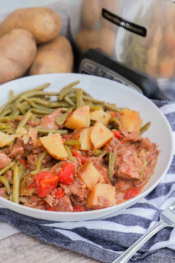 Slow Cooker Steak and Potatoes in a white bowl with slow cooker and potatoes in back