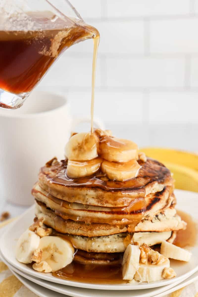 Banana Pancakes stacked on white plates with syrup pouring over top