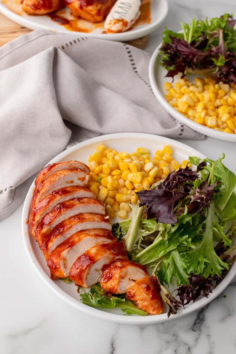 baked BBQ chicken sliced on white place with corn and salad.