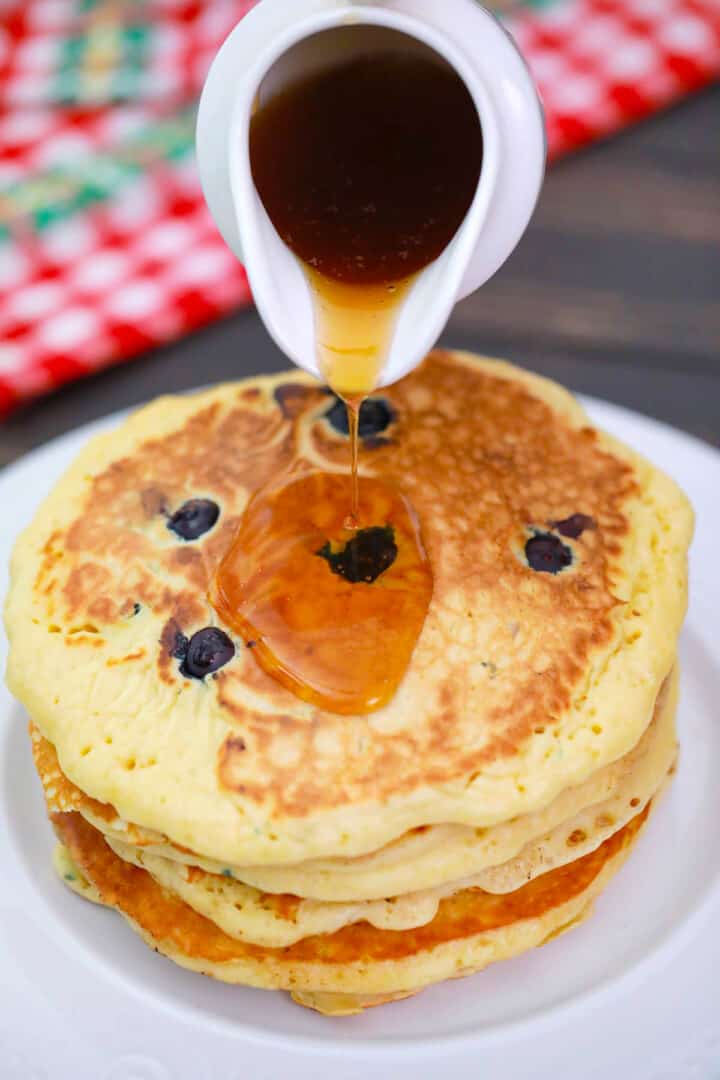 Blueberry Pancakes stacked on white plate pouring syrup on top.