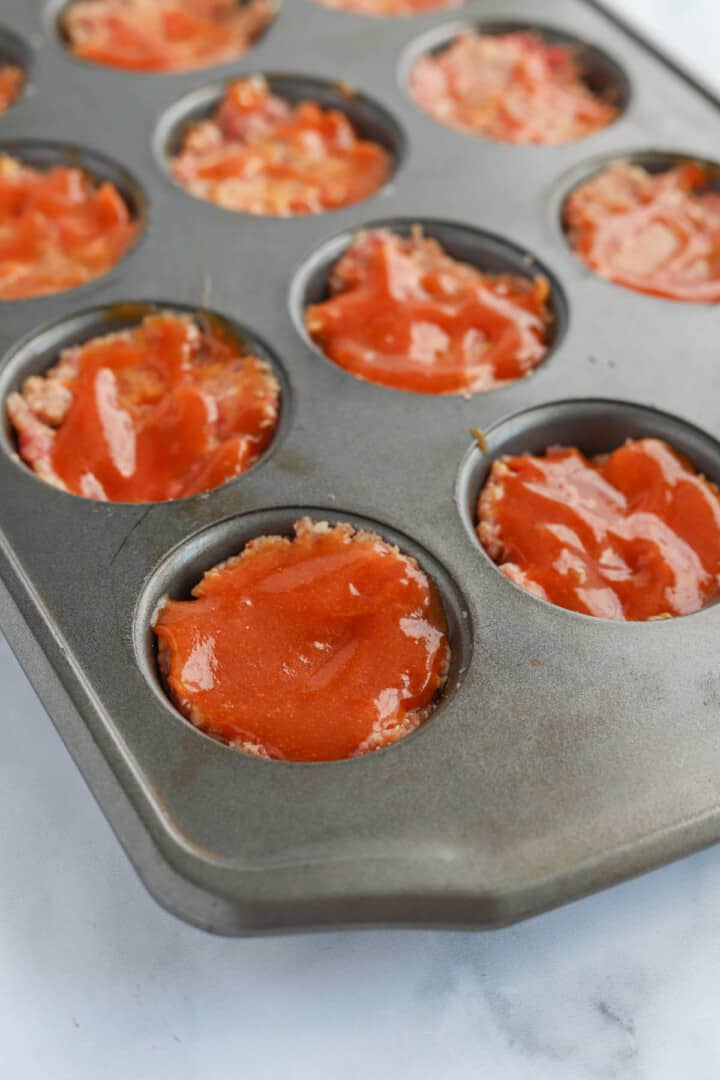 adding meatloaf mix to muffin tin and topping with sauce.