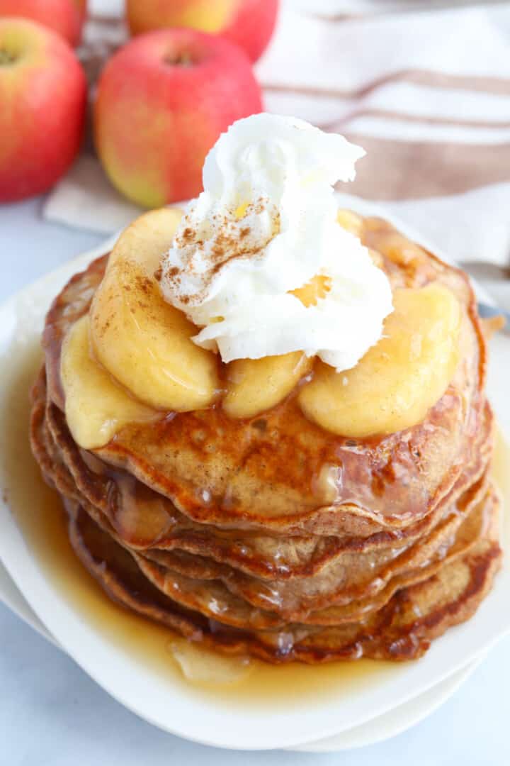 Apple Pie Pancakes stacked on white plate topped with whipped cream.