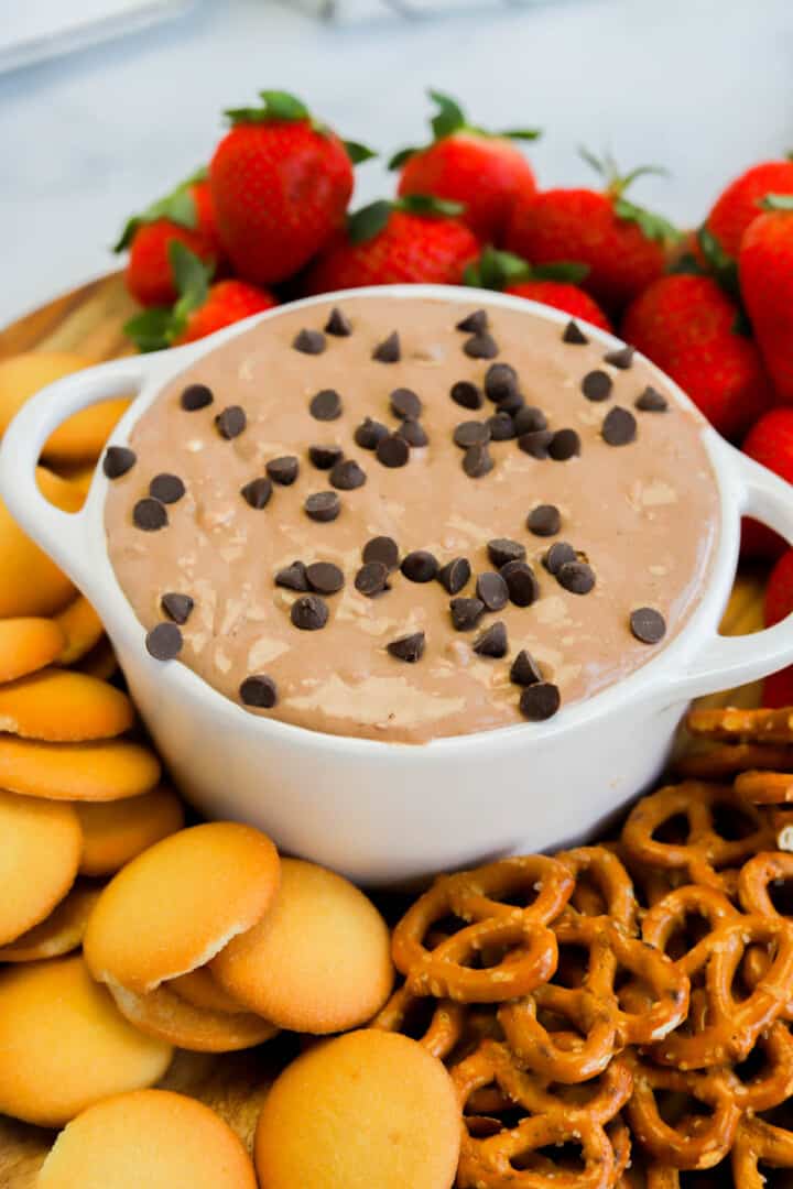 Brownie Batter Dip in white serving bowl surrounded by vanilla wafers, strawberries and pretzels.