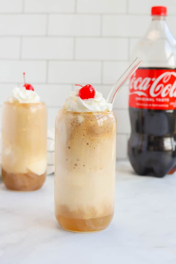 2 Coke Floats in glasses topped with whipped cream and cherry.
