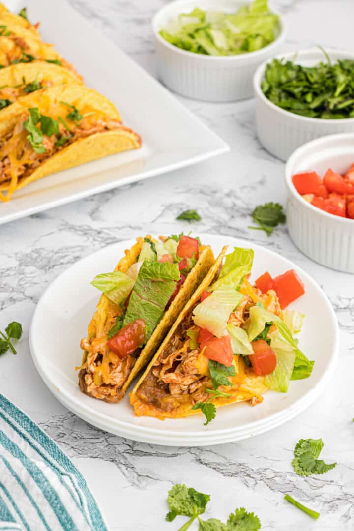 Baked chicken tacos on white plates with lettuce and diced tomatoes.