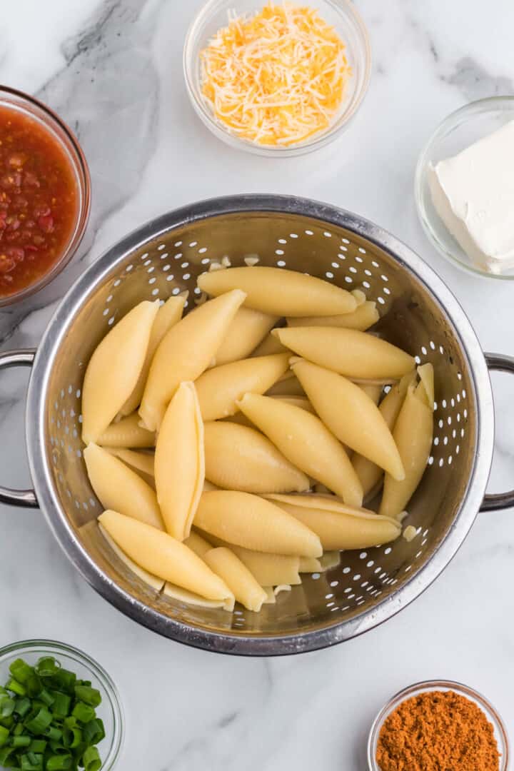 cooked pasta shells in colander with ingredients around.