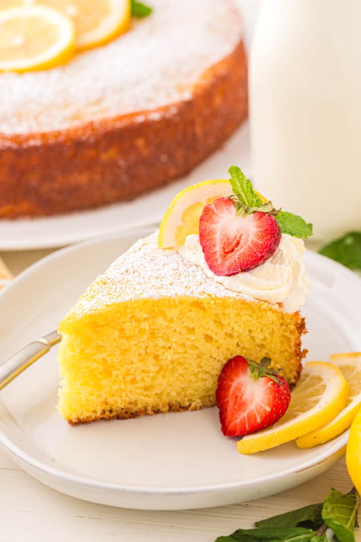 slice of lemon ricotta cake on white plate with lemon slices, whipped cream, and a strawberry slice.