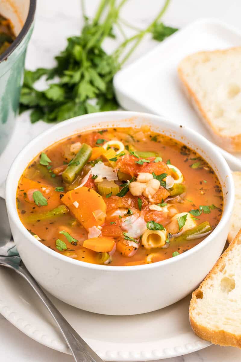 closeup of minestrone in white bowl with bread on the side.