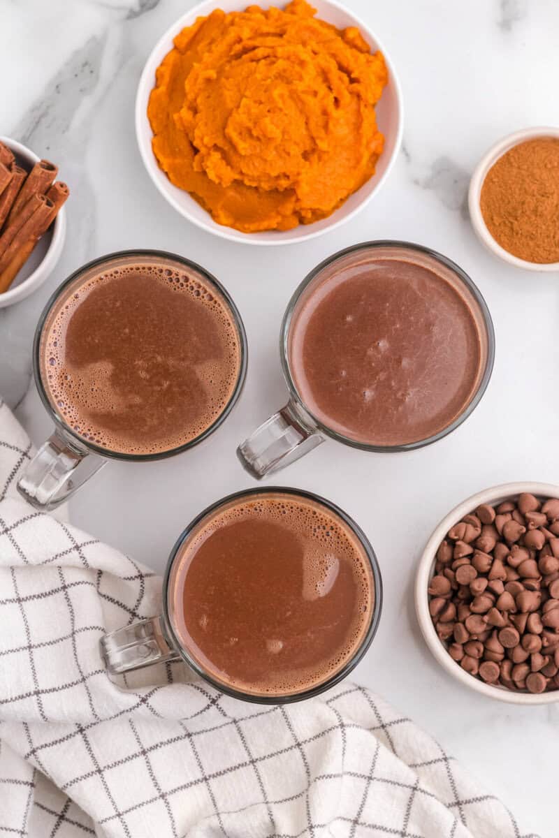 3 mugs of hot chocolate with ingredients around them.