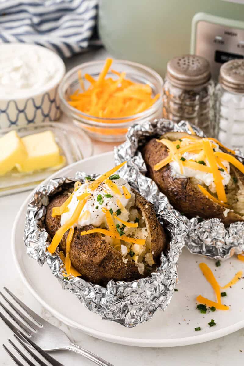 baked potatoes wrapped in foil on white plate topped with cheese and sour cream.