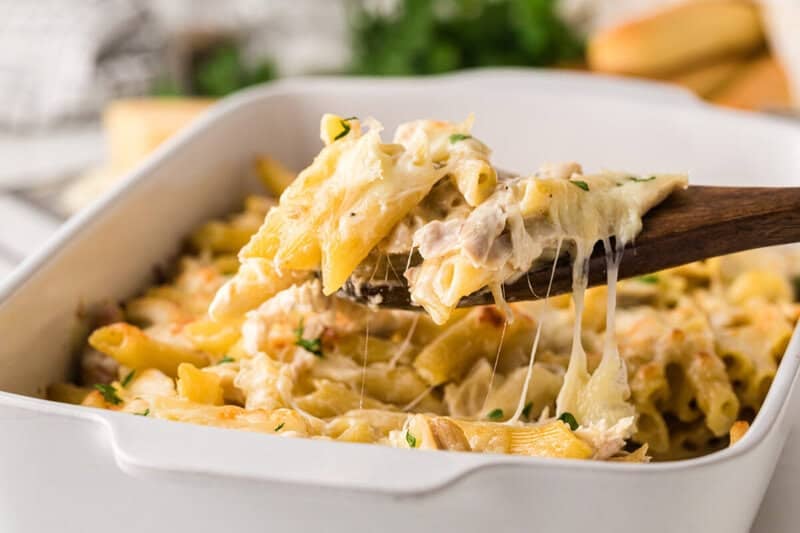 scooping chicken alfredo casserole bake from the baking dish with a wooden spoon