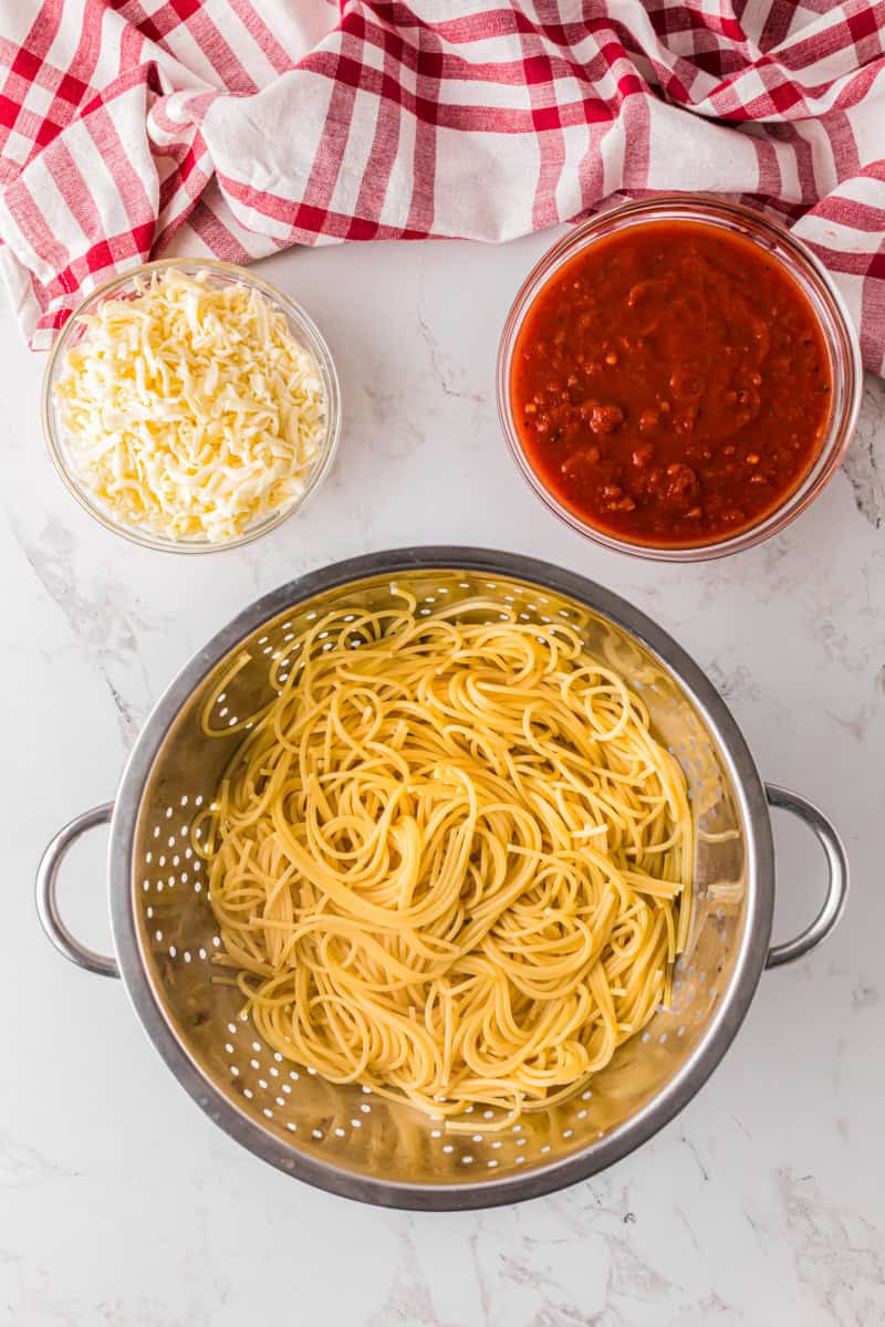 cooked spaghetti in the colander with the sauce and cheese next to it.