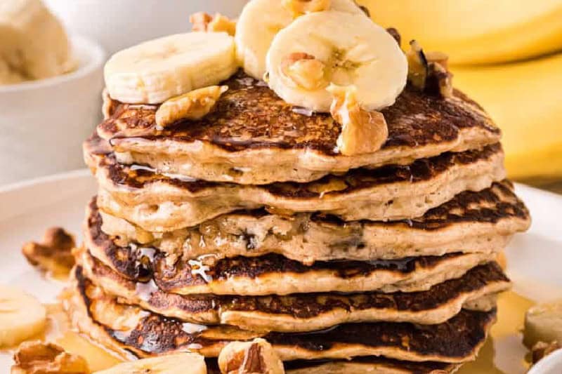 Stack of Banana Pancakes served on a white plate with slices of bananas and walnuts.