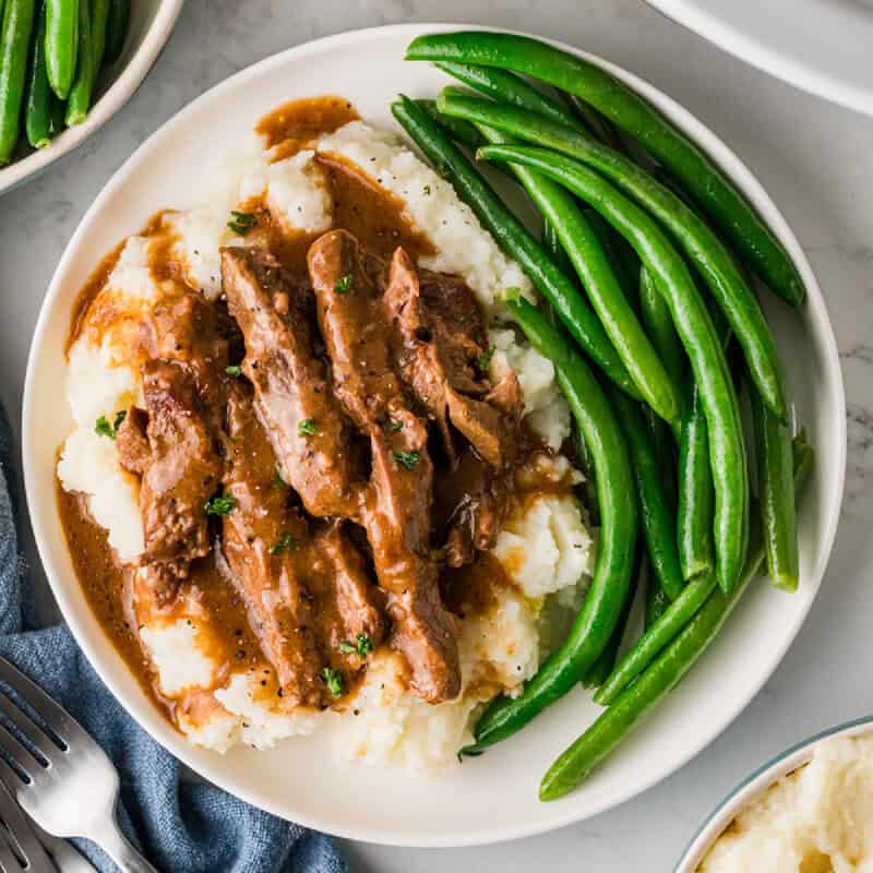 the slow cooker steak and gravy served on a bed of mashed potatoes and green beans.