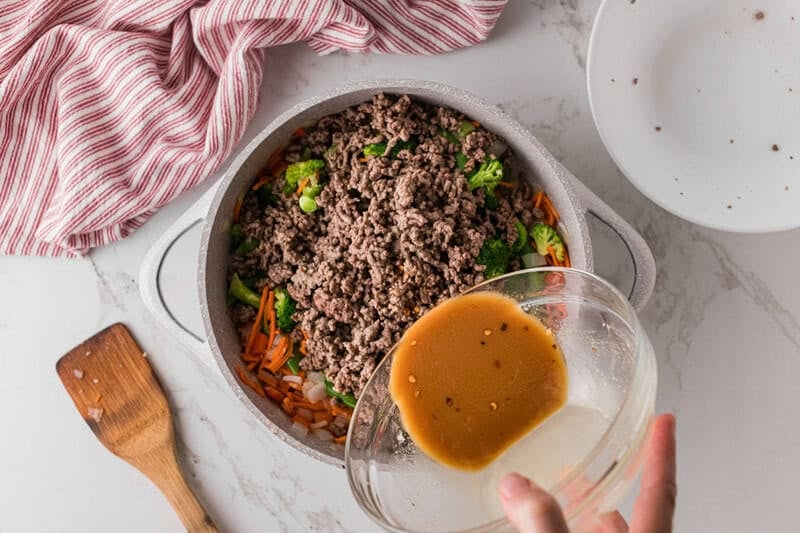 pouring the sauce over the ground beef stir fry in the pot.